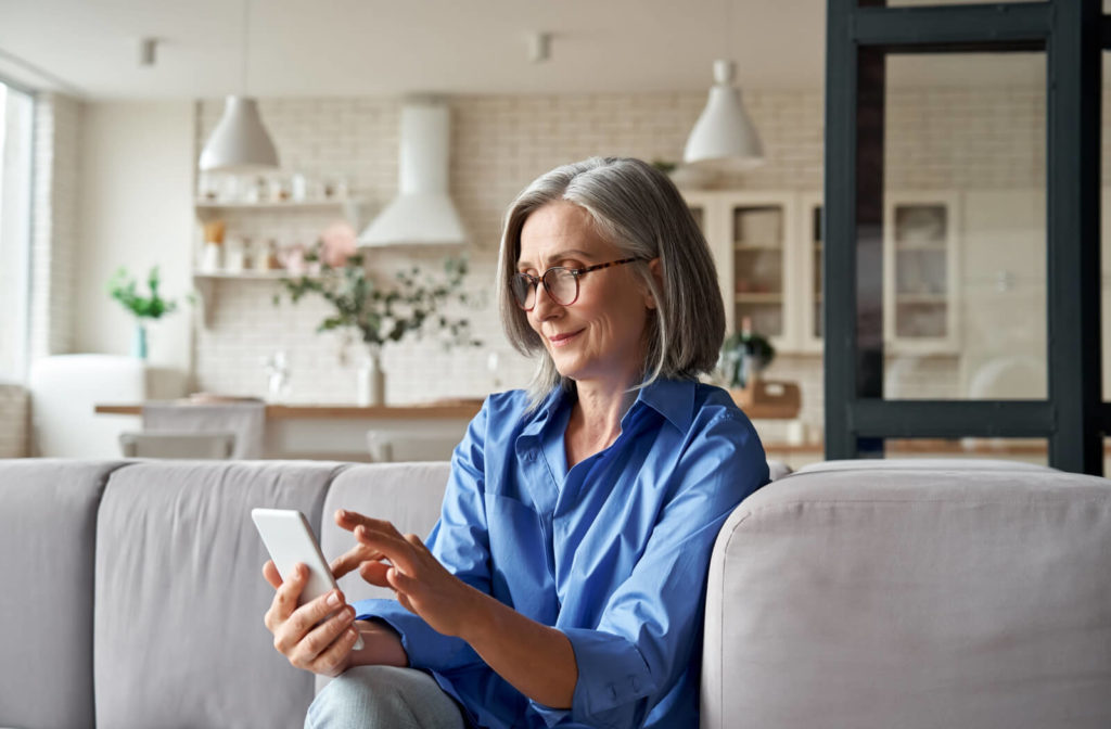 A senior woman with grey hair and glasses smiling while looking at her smartphone.