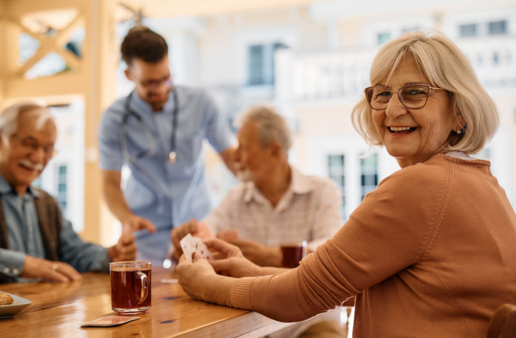 A senior woman sits at a table, holding cards next to a cup of tea and smiling. In the background, fellow seniors engage in conversation while a nurse helps out