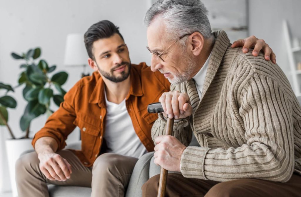 An adult child sits next to their older parent on the couch, a hand on their back, to broach the topic of assisted living