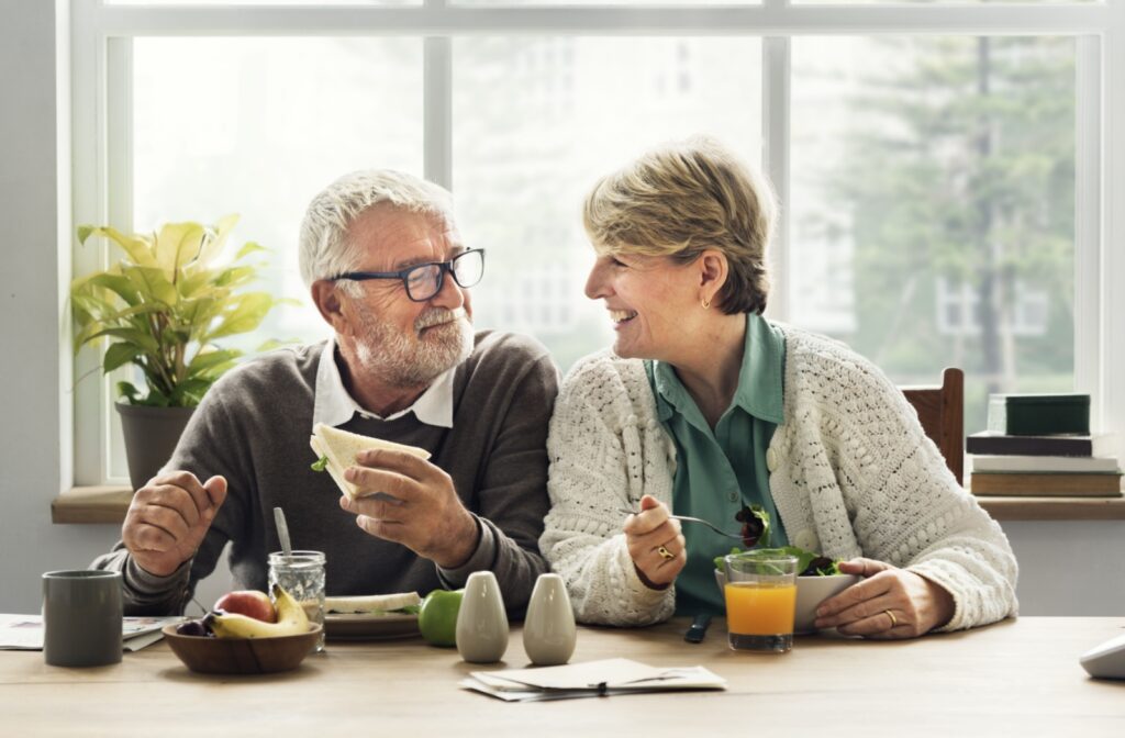 An older married couple smiling at each other while eating a brain-friendly breakfast in their kitchen.