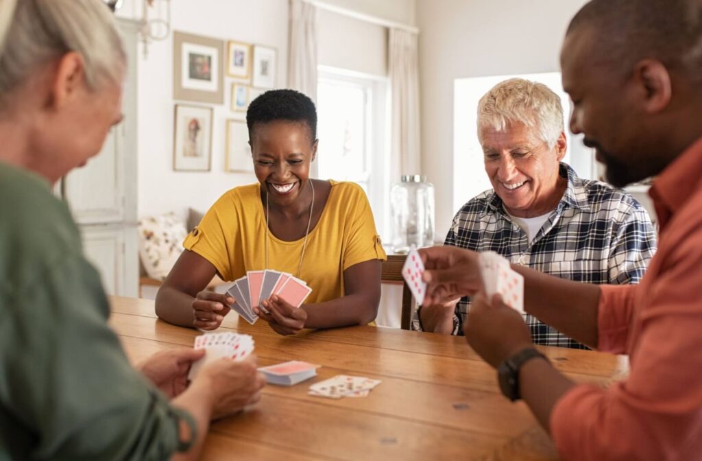 A group of older adults laugh while playing cards together in a brightly lit communal space in a senior living community