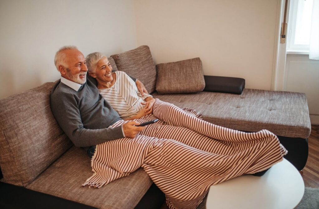 A senior couple cuddles underneath a blanket on a lounging couch while watching a movie about Alzheimer’s in the afternoon