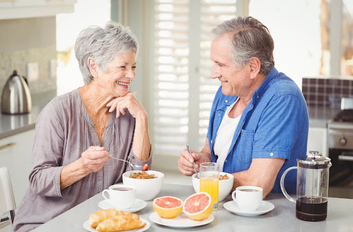 a senior couple smile at each other as they sit at a table eating bowls of oatmeal, coffee, grapefruit and coffee.