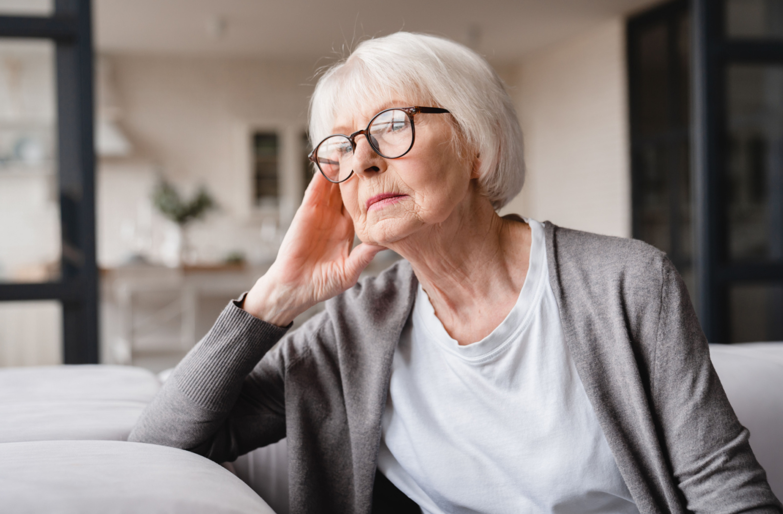 A senior woman with glasses sitting on a couch appears to be sad and looking outside the window