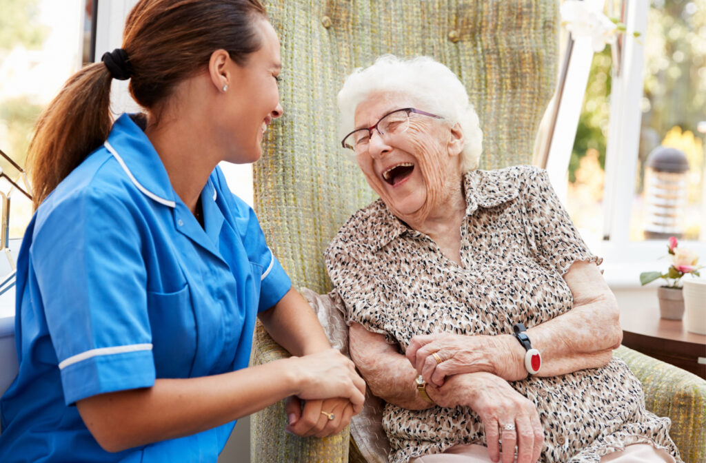 A senior woman sitting in a chair and talking to a caregiver in senior living community.