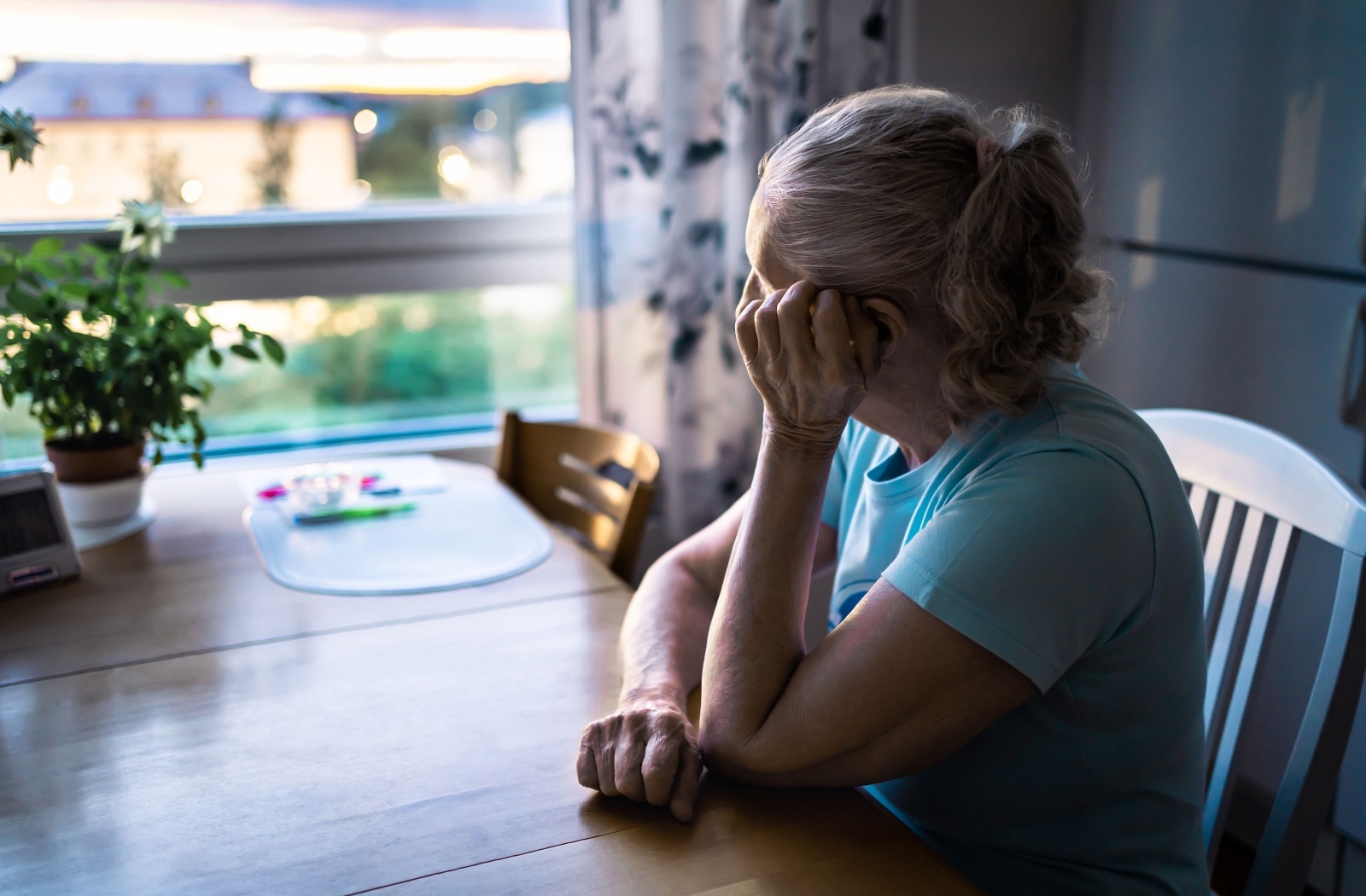 A lonely senior rests their head on their hand while they stare out their kitchen window.