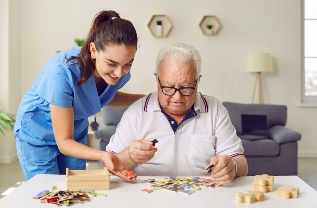 An elderly man focused on a cognitive game while a nurse stands beside him, offering encouragement in a memory care community.