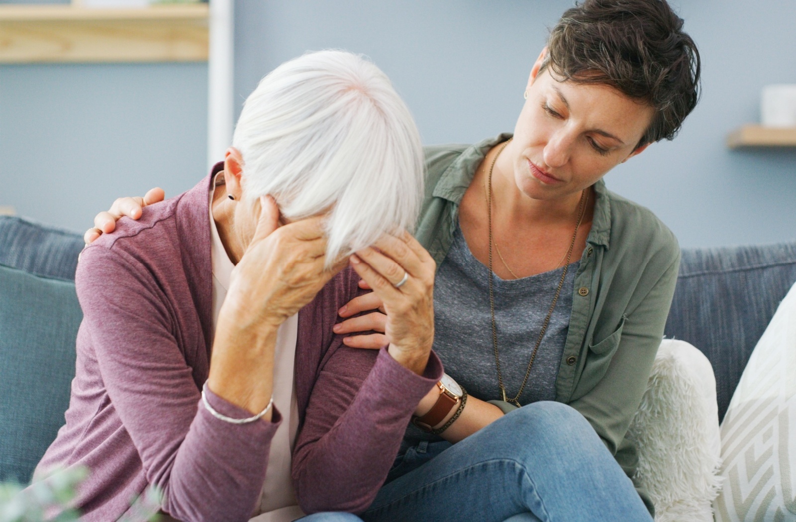 A senior with their head in their hands as if they are saddened by their dementia, with a younger family member next to them trying to comfort them.