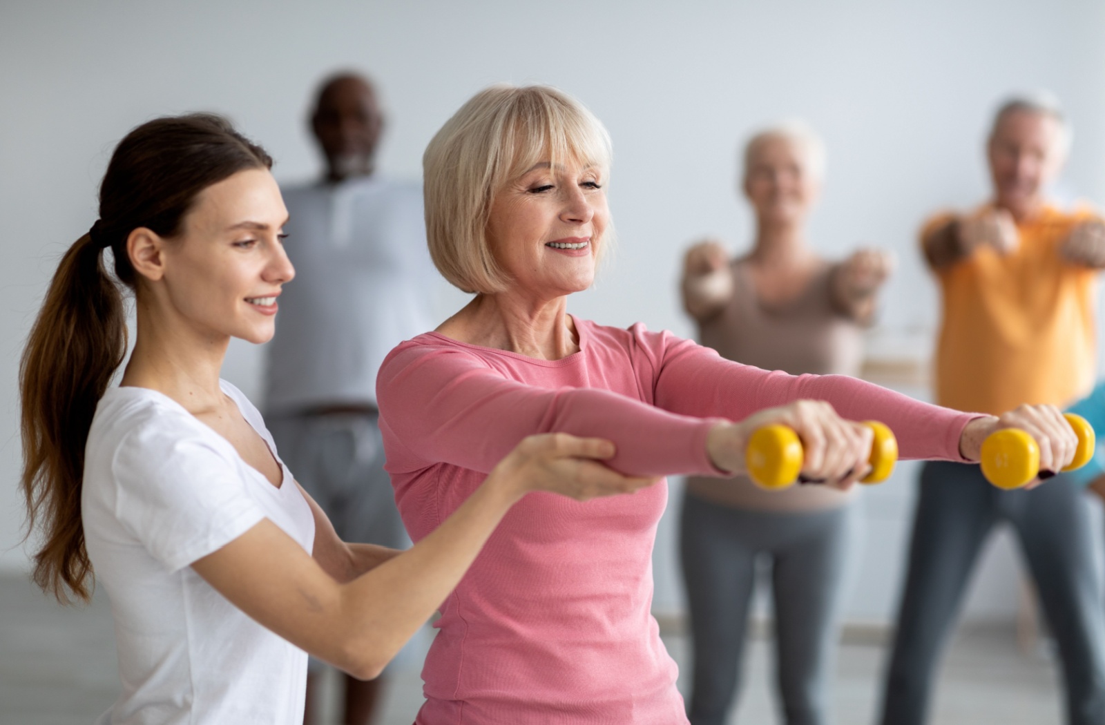 An assisted living staff member supporting a resident as they lift light weights in a group fitness class.