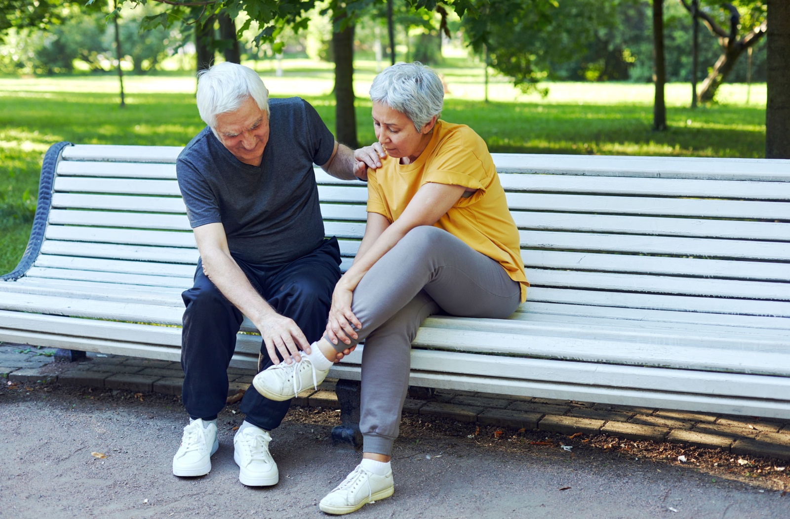 A senior man and woman sitting on a park bench as the woman holds her injured ankle.
