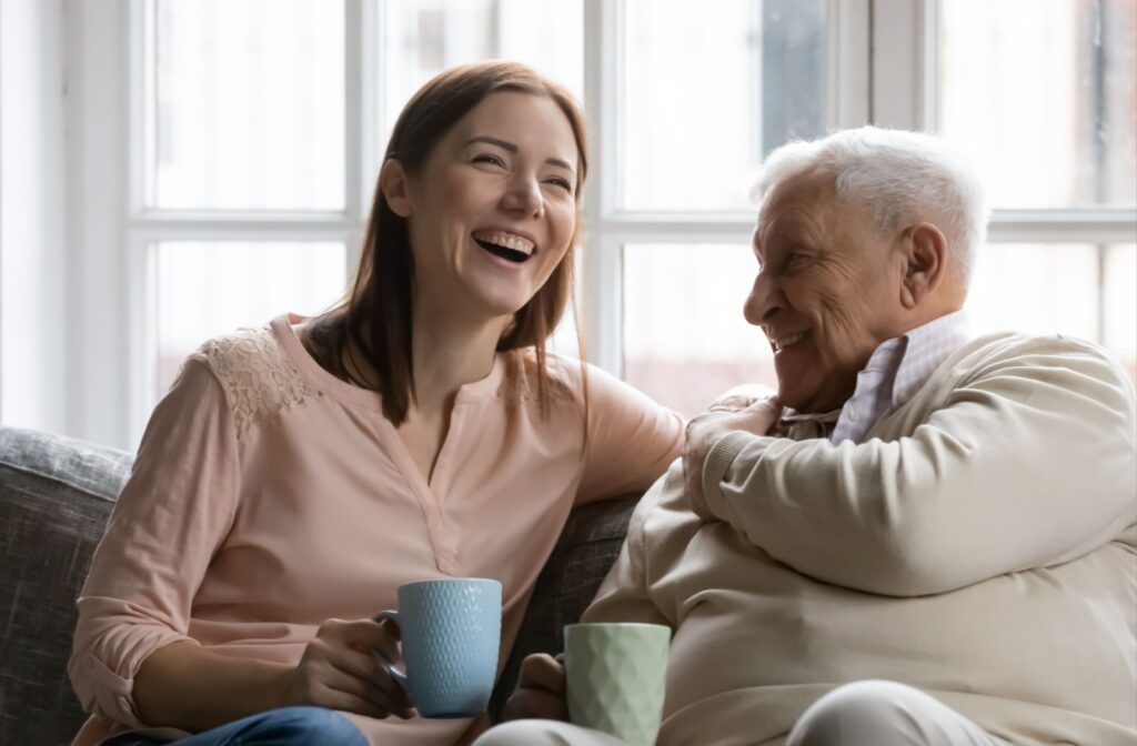 A senior sitting on a couch with a family member, laughing while holding a cup of tea or coffee.