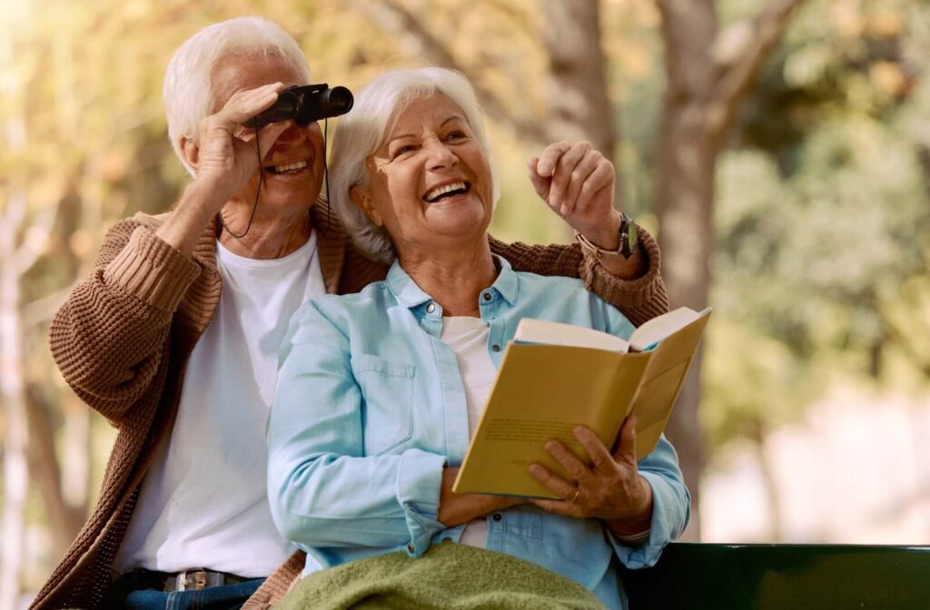 Two older adults sitting on a bench smiling while one looks through binoculars and the other reads a book outdoors