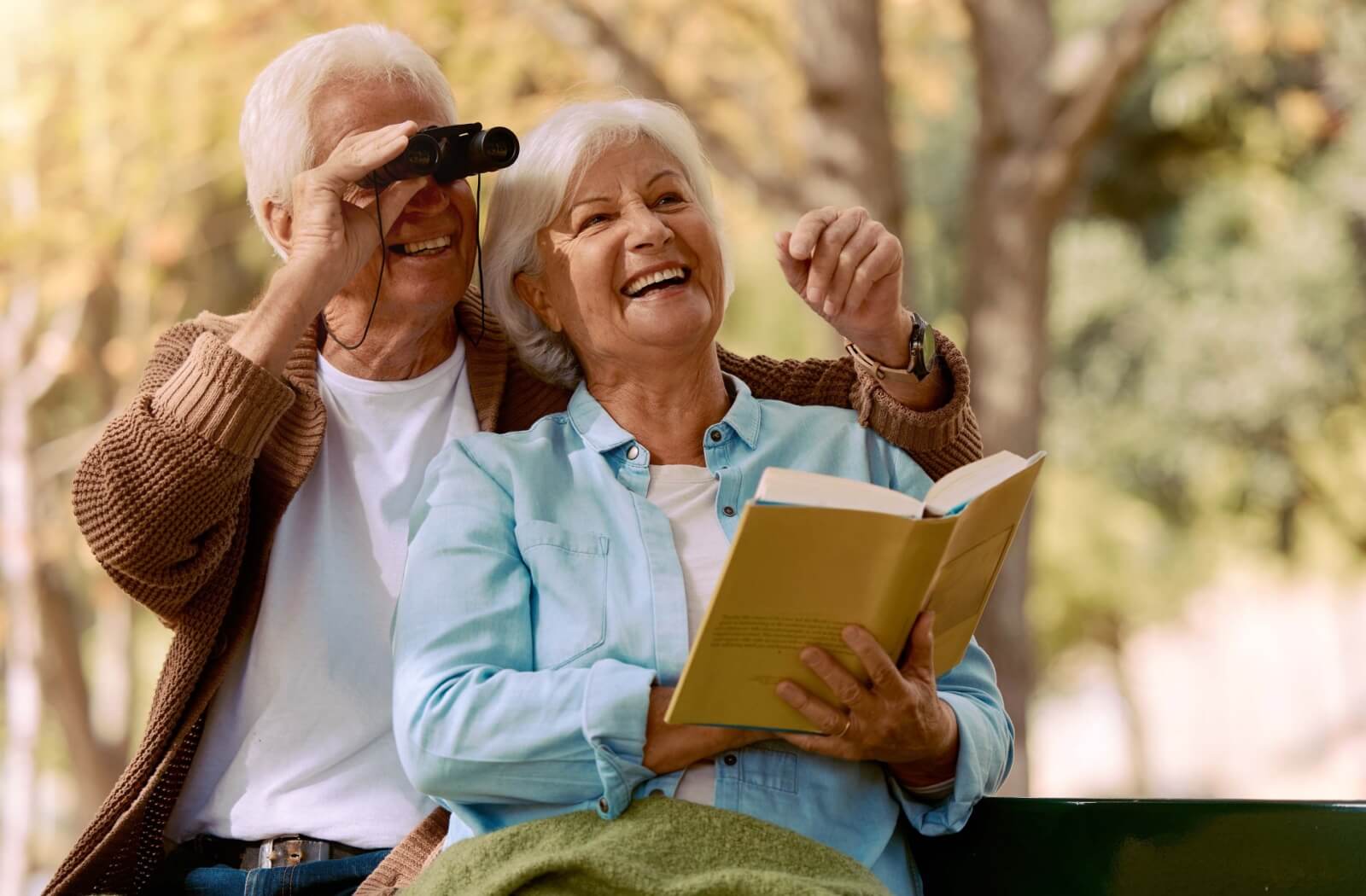 Two older adults sitting on a bench smiling while one looks through binoculars and the other reads a book outdoors