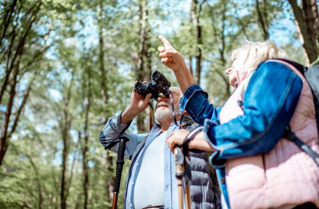 Two older adults birdwatching in the forest using binoculars and walking sticks while pointing at something in the trees