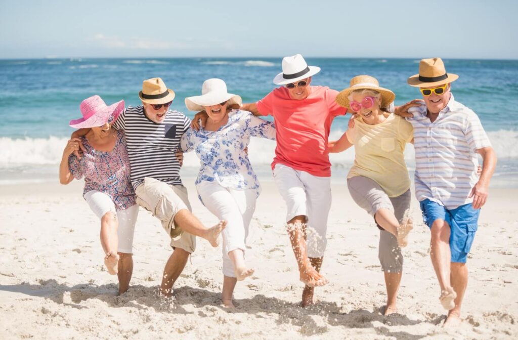 A group of seniors wearing large hats and sunglasses link arms to kick up sand at the beach, enjoying each other’s company