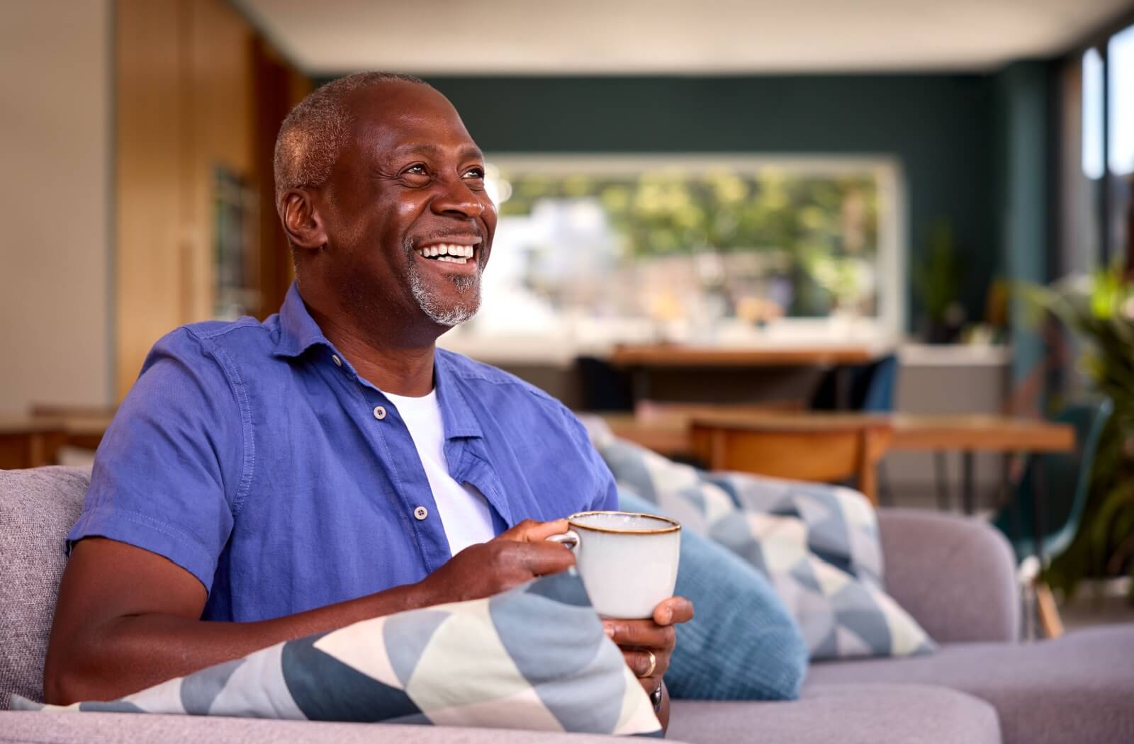 A happy senior smiles while enjoying a morning cup of coffee, relaxing on the couch
