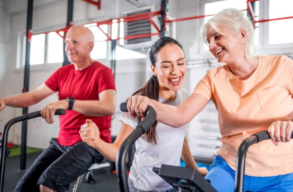 Two seniors in a gym use stationary bikes guided by a trainer who celebrates their progress